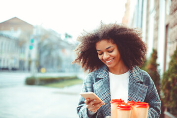 Young happy african american girl holding take away coffee cups and texting on smartphone outdoors,