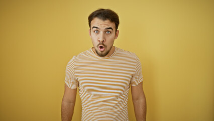 Surprised young man with beard against a yellow wall wearing striped shirt showing expressive emotion.