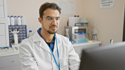 A young hispanic man with a beard, wearing glasses and a lab coat, works attentively in a hospital...