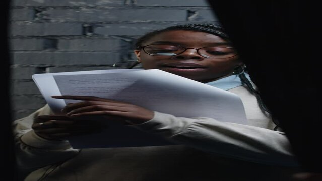 Vertical medium shot of young Black woman revising script before auditions in theater, standing against brick wall under projector light