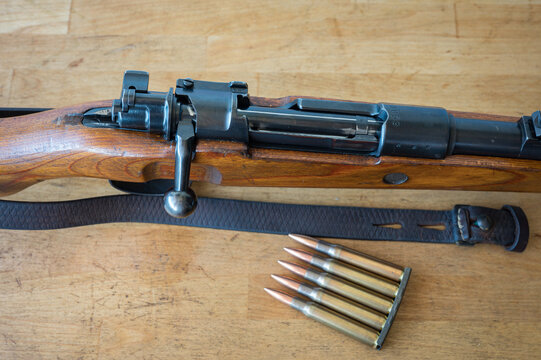 Detail of an ammunition clip with 5 30-06 caliber rifle bullets next to a Mauser-type bolt-action military rifle on a wooden table