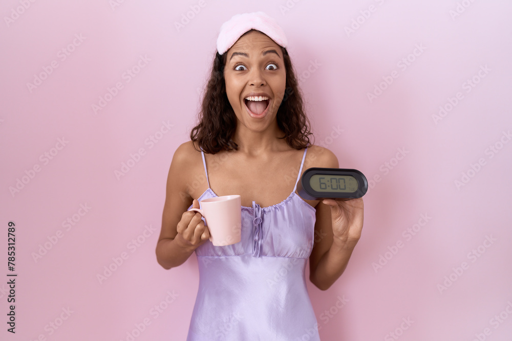 Poster Young hispanic woman wearing nightgown holding alarm clock celebrating crazy and amazed for success with open eyes screaming excited.