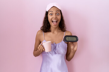 Young hispanic woman wearing nightgown holding alarm clock celebrating crazy and amazed for success...