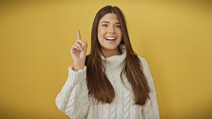 A cheerful young hispanic woman in a white sweater posing with an idea gesture isolated against a...