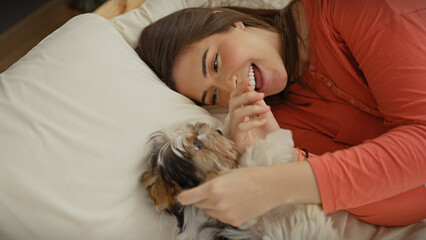 Hispanic woman laughing with a biewer yorkshire terrier on a bed, showcasing joy and companionship indoors. - obrazy, fototapety, plakaty