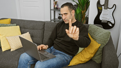 Hispanic man giving disapproval sign while working on laptop in a modern living room. - obrazy, fototapety, plakaty