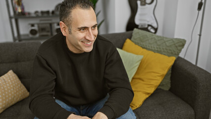 Smiling man sitting comfortably on a gray sofa in a cozy living room with colorful pillows and a guitar background - Powered by Adobe