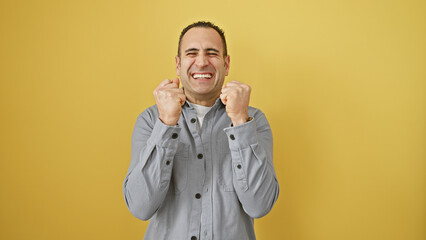 A joyful hispanic man celebrating with clenched fists against a vibrant yellow isolated background.