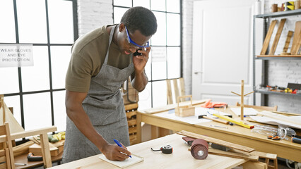 A young man works attentively in a workshop, wearing safety glasses and an apron while taking notes...