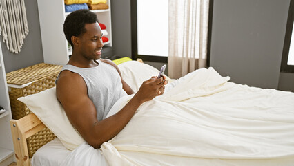 A young african american man in a tank top using a smartphone while relaxing in a bedroom with white bedding. - Powered by Adobe