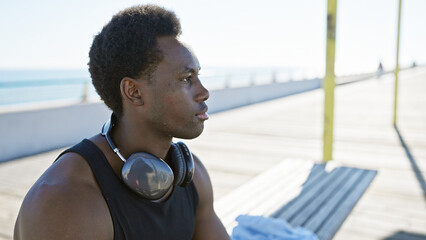 A young african american man with headphones gazes thoughtfully by the seaside promenade under the...