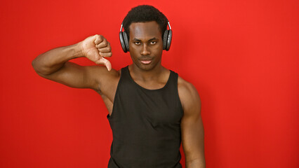 A young african american man with headphones showing thumbs down against a red isolated background.
