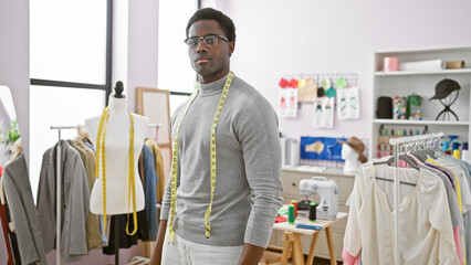 Confident man with glasses and measuring tape in a modern atelier with mannequin and clothing rack.