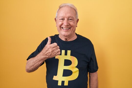 Senior man with grey hair wearing bitcoin t shirt doing happy thumbs up gesture with hand. approving expression looking at the camera showing success.