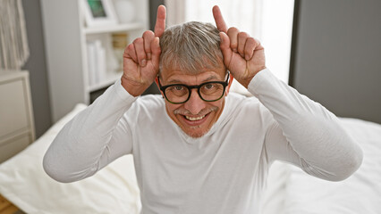 Smiling middle-aged man with gray hair making bunny ears gesture while sitting on a white bed in a...