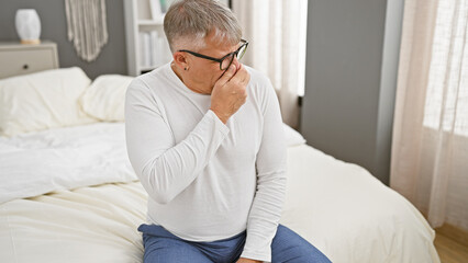 Middle-aged man pinches nose to avoid bad odor while sitting on bed in a well-lit bedroom