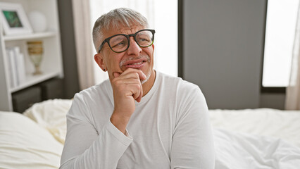 A thoughtful middle-aged man with grey hair wearing glasses sits on a bed indoors, hand on chin,...