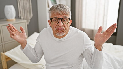 Confused senior man with grey hair and glasses sitting on a bed in a bedroom raises his hands in a...