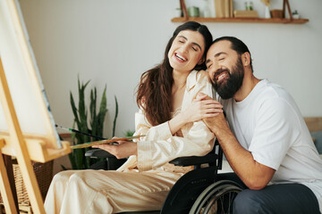attractive woman with mobility disability painting on easel next to her cheerful bearded husband