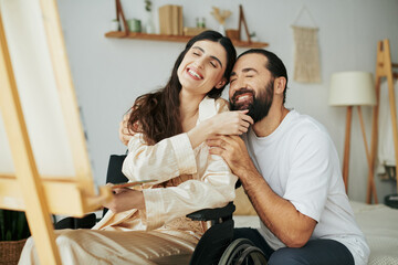 attractive woman with mobility disability painting on easel next to her cheerful bearded husband - obrazy, fototapety, plakaty