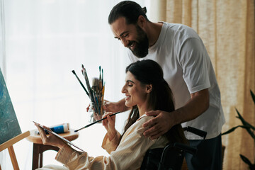 caring jolly husband helping his inclusive good looking wife to paint on easel while at home - obrazy, fototapety, plakaty