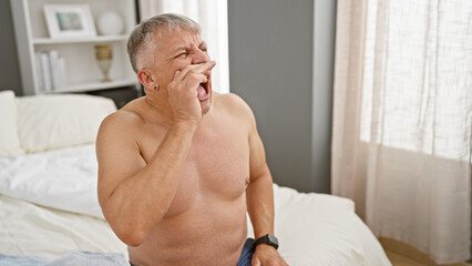 Shirtless senior man with grey hair yawning while sitting on a bed in a bedroom.