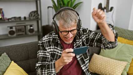 Excited senior man with headphones playing smartphone game on couch at home