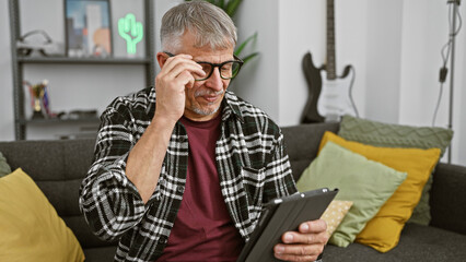 A grey-haired man adjusts his glasses while reading a tablet in a stylish living room.