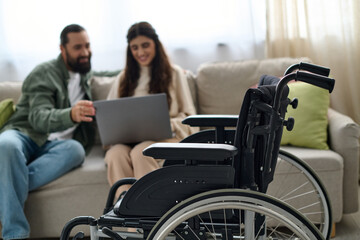 focus on modern wheelchair in front of blurred bearded man and disabled woman looking at laptop - obrazy, fototapety, plakaty