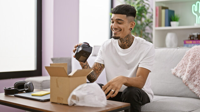 Joyful young latin man, grinning ear to ear, unboxing his new professional camera at his comfy home