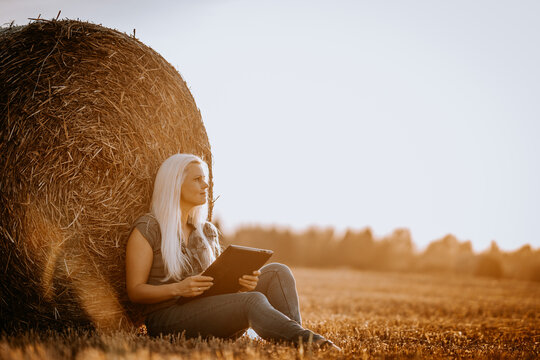 Valmiera, Latvia - August 17, 2024 - A contemplative woman sits by a hay bale in a field with a tablet, looking off into the distance during sunset.