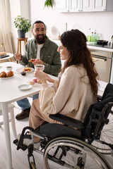 joyful woman with inclusivity in wheelchair eating sweets at breakfast with her handsome husband