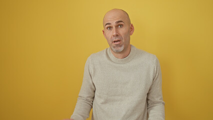 A perplexed bald man with a beard wearing a sweater standing against a yellow background.