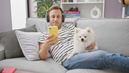 At home, a young man, casually engrossed in the digital world, sits back relaxed on the sofa with his faithful dog. he is deeply lost in his own world of sound, using his smartphone and headphones.