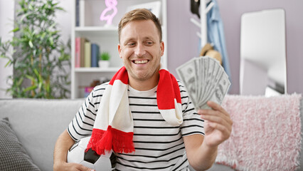 Excited young caucasian man clutching dollars, holding soccer ball in home interior, passionate...
