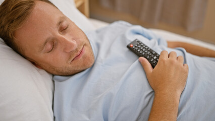 Exhausted young caucasian man, remote in hand, indulges in comfortable sleep on cozy bed, holding...