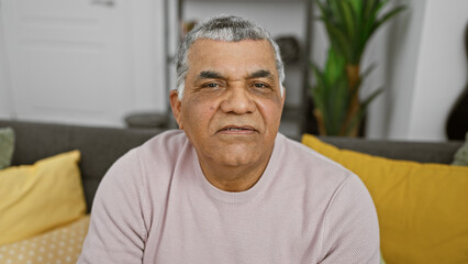 Obraz premium Handsome senior latin man with grey hair posing in a cozy living room setting, exuding maturity and relaxation at home.