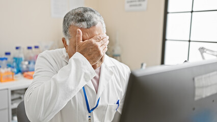 A stressed senior man in a lab coat covers his face with his hand in an indoor laboratory setting.