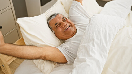 A joyful senior man with grey hair relaxing in a bright bedroom, stretching his arms with a smile