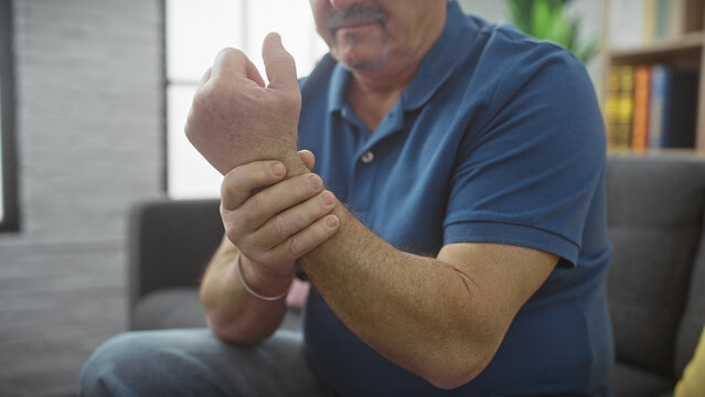 Fototapeta A mature man in a blue shirt expressing discomfort while holding his wrist at home, suggesting pain or injury.