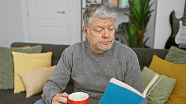 A mature man relaxes at home with coffee and a book on a cozy couch adorned with colorful pillows.