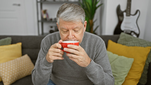 A grey-haired man enjoys a cup of coffee in a cozy living room with colorful pillows and a guitar in the background.