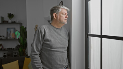 A pensive grey-haired man gazes out a window in a modern room