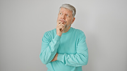A pensive senior man with grey hair in a turquoise sweater stands against a white background, hand...