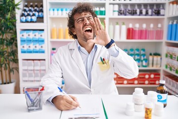 Hispanic young man working at pharmacy drugstore shouting and screaming loud to side with hand on...