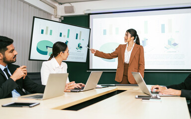 A cheerful and confident Asian businesswoman stands, present bar charts data from projector screen to her office colleagues. Asian business women leader role at the meeting. - 785326990