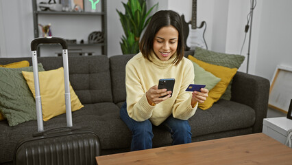 Smiling hispanic woman booking travel online using smartphone and credit card in living room with...