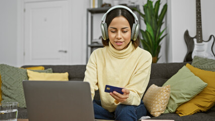A young hispanic woman wearing headphones uses a credit card for online shopping on a laptop in a cozy living room.