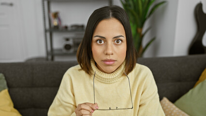 Portrait of a young hispanic woman with glasses in a yellow sweater, sitting in a modern living...