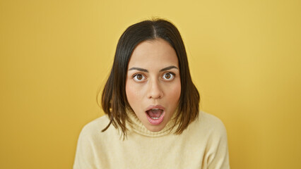 A surprised young hispanic woman with an open-mouth expression stands against a vibrant yellow...